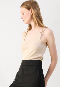 Beige knit top with wide straps featuring chain accents, side seams, and a scoop neckline, paired with black high-waisted trousers.