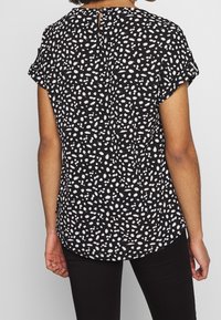 Czarna bluzka z białym wzorem w kropki, krótkimi rękawami, zaokrąglonym dołem oraz wycięciem w kształcie klucza z tyłu przy szyi. Miękka tekstura tkaniny.