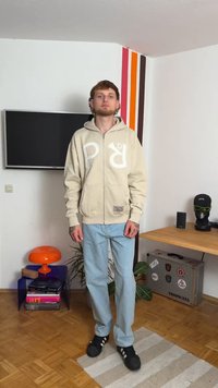 Cream zip-up hoodie with large white logo, paired with light blue straight-leg jeans and black sneakers. Background includes a TV and orange lamp.