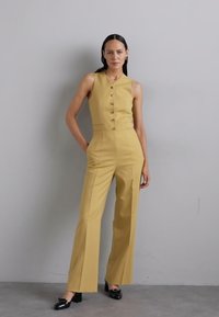 Tutina gialla con un corpetto aderente, scollo a V e bottoni frontali. Taglio a gamba larga e tessuto morbido. Modella che indossa scarpe nere in vernice.
