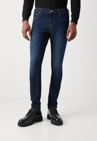 Jeans de mezclilla azul oscuro, con corte ajustado, diseño de cinco bolsillos y costuras naranjas en contraste. Llevados con zapatos de cuero negros.