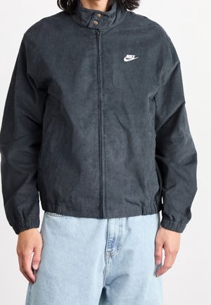 Mężczyzna ubrany w ciemnoszarą kurtkę Nike na suwak z wysokim kołnierzem i jasnoniebieskie dżinsy, stoi na tle jednolitej białej ściany.