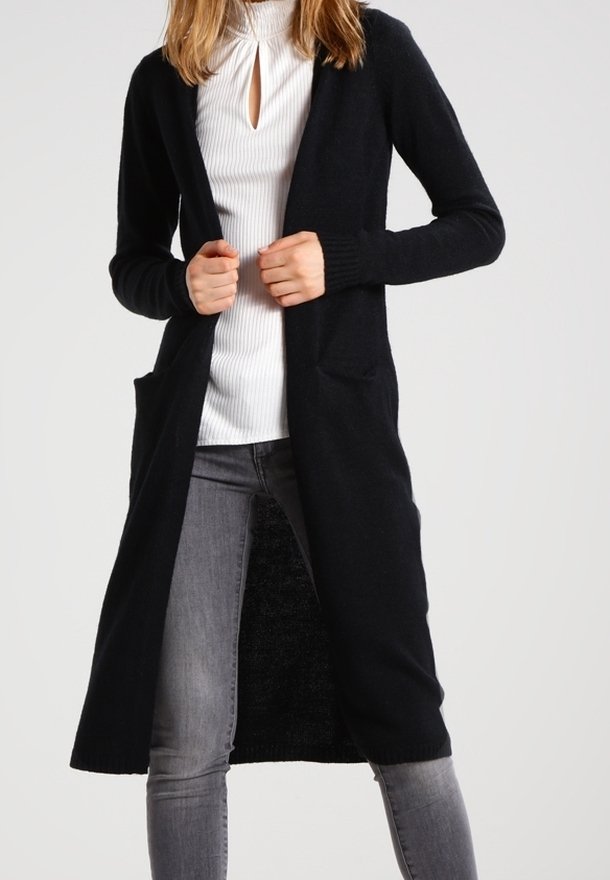 Femme portant un long cardigan noir ouvert sur le devant, par-dessus un top blanc côtelé avec un décolleté en trou de serrure, et un jean skinny gris, debout devant un fond uni.