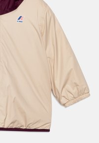 Giacca leggera beige con colletto bordeaux. Presenta polsini elastici e un logo K-Way, realizzata in materiale liscio e resistente all'acqua.