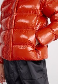 Giacca a vento rosso lucido con sezioni imbottite a trapunta e una tasca laterale con zip. Indossata sopra pantaloni neri larghi.