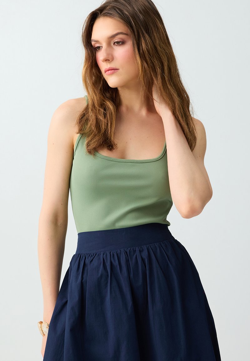 Jeune femme aux cheveux bruns mi-longs portant un haut vert sans manches et une jupe bleu marine taille haute, main touchant le cou, regard baissé.