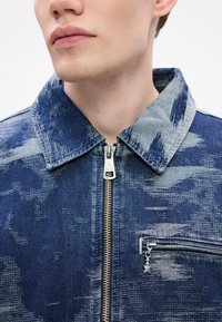 Chaqueta de denim con un patrón texturizado en azul, cuello en contraste, cierre frontal de cremallera, y un pequeño bolsillo en el pecho con un charm en forma de estrella plateada.