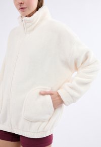 Bílá fleece bunda s vysokým límcem, zipem vpředu a velkými zakřivenými kapsami. Materiál má jemnou, texturovanou úpravu.