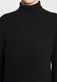 Pull à col roulé en tricot côtelé noir avec un motif en V subtil sur la poitrine, porté par une personne visible du cou jusqu'au milieu du torse.