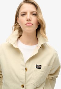 Chemise en velours côtelé beige avec un col rabattu, une patte de boutonnage et une poche poitrine ornée d'un patch logo. Tissu texturé avec des côtes visibles.