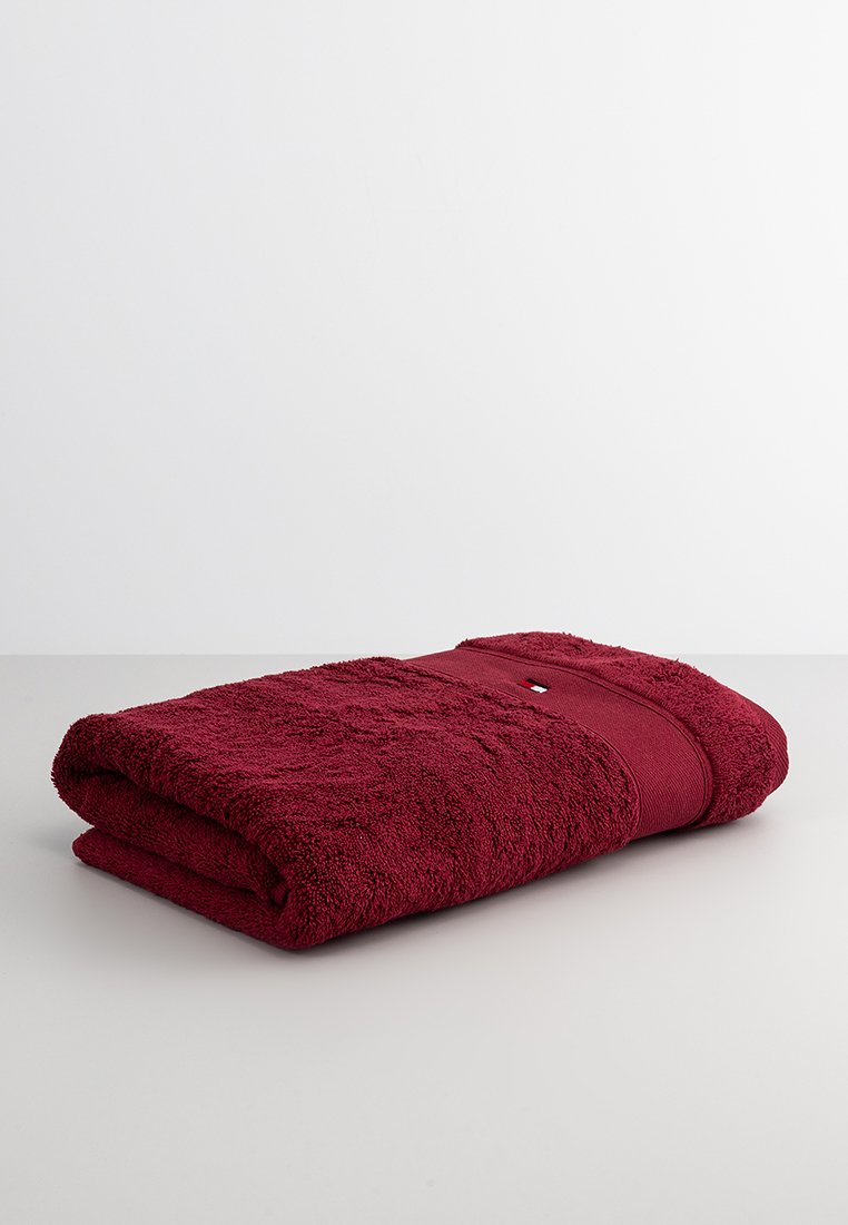 Asciugamano in cotone bordeaux, texture soffice, spessore spesso con una fascia a righe, caratterizzato da un piccolo dettaglio con logo sul bordo, piegato con cura.