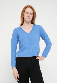 Jauna sieviete ar sarkaniem matiem, tērpusies zilā V-veida kakla džemperī un melnās biksēs, stāv ar vienu roku uz gurniem baltā fona.