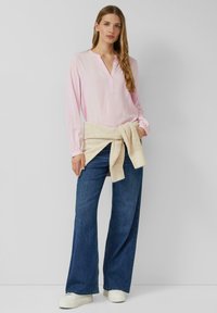 Jasnoróżowa bluzka z długim rękawem i zapiętym na guziki kołnierzykiem, zestawiona z szerokimi niebieskimi jeansami. Beżowy dziany sweter jest przewiązany w talii.