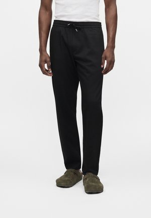 Homme portant un pantalon noir à cordon, une chemise blanche et des chaussures à enfiler vert olive, debout sur un fond uni.