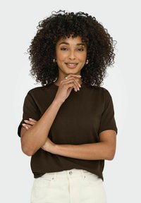 Maglietta a maniche corte marrone con vestibilità rilassata, realizzata in materiale morbido. Il modello posa con le braccia incrociate, indossando pantaloni color crema.