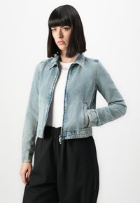 Ženska s ravnimi temnimi lasmi, oblečena v svetlo modro jeans jakno preko bele srajce in ohlapnih črnih hlač, stoji pred belo ozadjem.