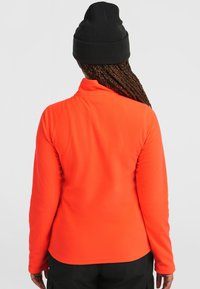 Sudadera de polar naranja para mujer con cuello alto y mangas largas, mostrada desde atrás. Textura suave y acabado liso. Se lleva un gorro negro.