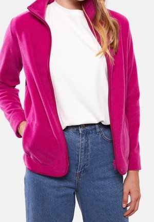 Kvinde iført en knaldpink fleecejakke med lynlås over en hvid t-shirt og blå denimjeans, stående med den ene hånd i lommen.