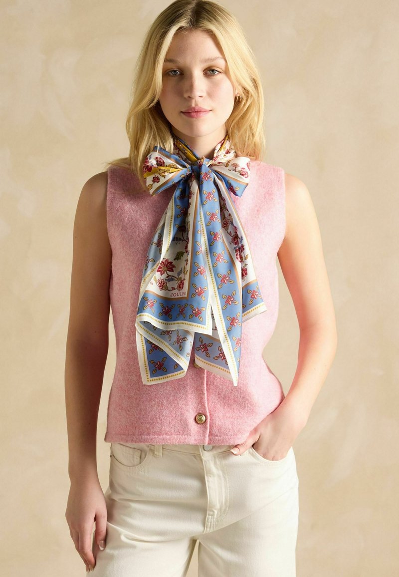 Vaaleanpunainen villainen hihaton paita kultaisella napilla, yhdistetty siniseen kukalliseen silkkihuiviin, joka on solmittu rusetille kaulassa. Vaaleat housut.