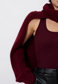 Persoon in een mouwloos bordeauxrood topje, zwarte leren broek en een bijpassende zachte, fuzzy bordeauxrode sweater over de schouders gedrapeerd.