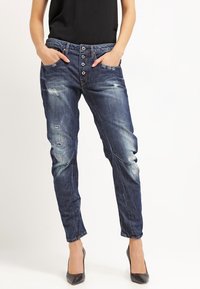 Jeans de mezclilla azul desgastado con un corte relajado, cierre de cinco botones, detalles desgastados y piernas entalladas, combinados con tacones negros.