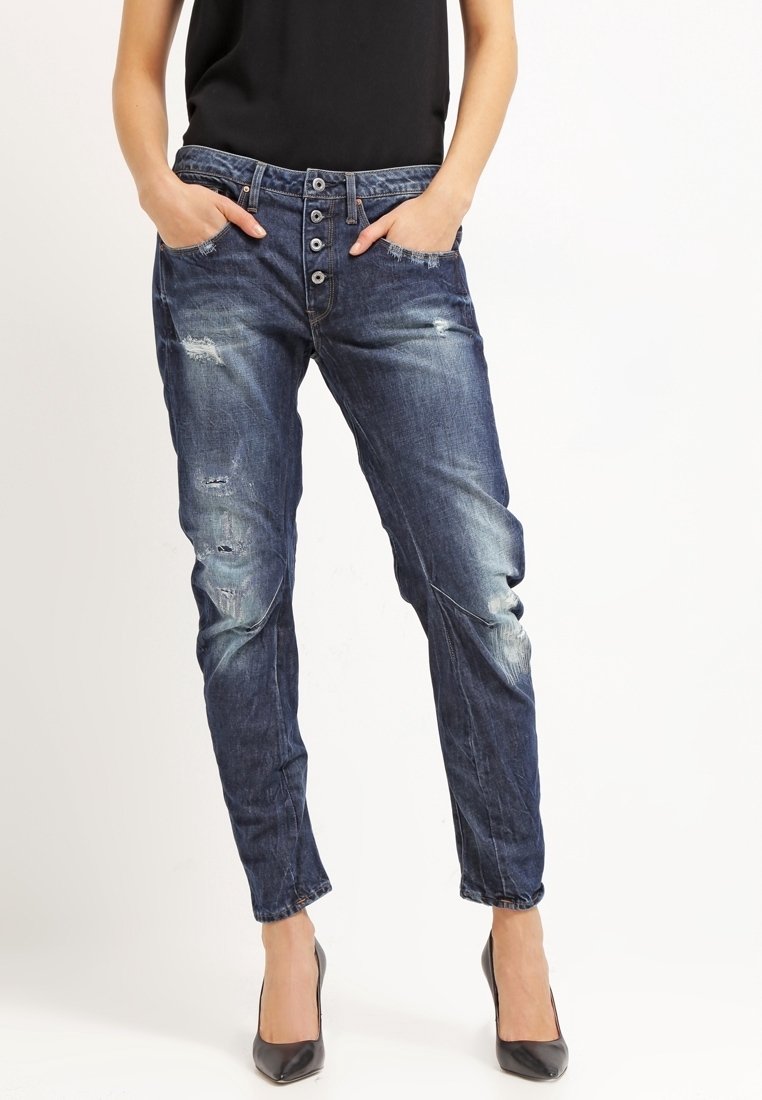 Jeans de mezclilla azul desgastado con un corte relajado, cierre de cinco botones, detalles desgastados y piernas entalladas, combinados con tacones negros.