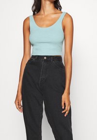 Donna che indossa un top corto senza maniche di colore azzurro chiaro e jeans neri a vita alta e larghi su uno sfondo semplice.