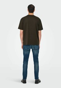 Mann steht mit dem Rücken zur Kamera, trägt ein dunkelbraunes kurzärmeliges T-Shirt, blaue Jeans und schwarze Schuhe vor einem schlichten hellen Hintergrund.