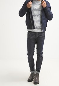 Marineblauer Bomberjacke über einem grau gestreiften Pullover, kombiniert mit dunklen, schmal zulaufenden Jeans und umgeschlagenen Ärmeln. Schwarze Knöchelstiefel runden das Outfit ab.