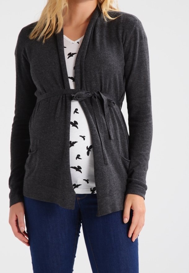 Femme portant un cardigan gris foncé noué par-dessus une chemise blanche avec des imprimés d'oiseaux noirs et un jean bleu, debout devant un fond uni.