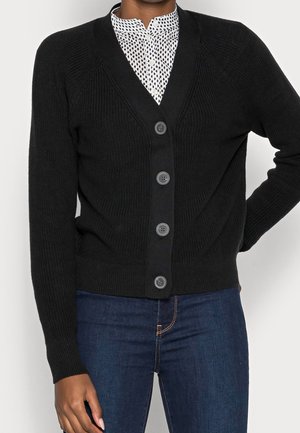 Black buttoned cardigan worn over white patterned blouse, paired with dark blue jeans, shown from shoulders to hips.