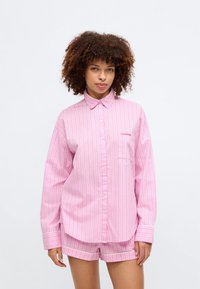 Ροζ καρώ πουκάμισο με κουμπιά, μακρύ μανίκι και τσέπη στο στήθος; συνδυασμένο με ροζ σορτς που έχουν λευκή περίμετρο.