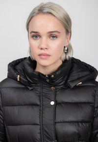 Zwarte pufferjack met capuchon, voorzien van een quilted ontwerp, gouden hardwareaccenten en knoopsluiting aan de voorkant.