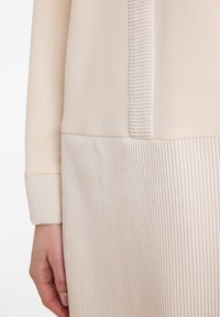 Langärmliges beiges Kleidungsstück mit strukturiertem, geripptem Stoff im unteren Bereich und glattem oberen Teil, mit gerippten Bündchen.