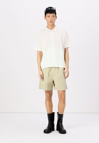 Polo creme de manga curta e textura, combinado com calções bege e botas de couro pretas até ao joelho. Óculos de sol a usar na cabeça.