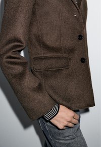 Brown wool blazer with a smooth texture, single-breasted design, and black buttons. Features a side pocket and a striped cuff.