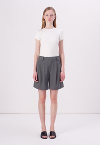 Hvit ribbet t-skjorte med korte ermer kombinert med grå, plisserte shorts. Modellen står og har på seg svarte sandaler. Enkel og minimalistisk design.
