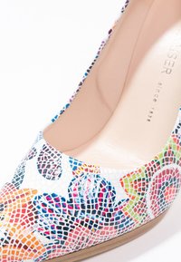 Gros plan sur un escarpin blanc à talon haut avec des motifs floraux mosaïques colorés et une semelle intérieure beige avec le texte de la marque.