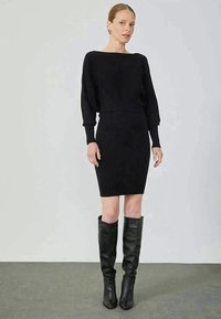 Vestido de malha preta canelada com decote barco, mangas compridas e silhueta justa; combinado com botas de couro pretas até ao joelho.