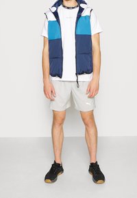 Chaleco acolchado en azul marino y turquesa con capucha sobre una camiseta blanca y pantalones cortos grises. Zapatillas deportivas negras con superficie texturizada y suela de goma.