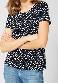 Černý krátký rukáv top s bílým puntíkovaným vzorem. Materiál má hladkou texturu, kulatý výstřih a volný střih.