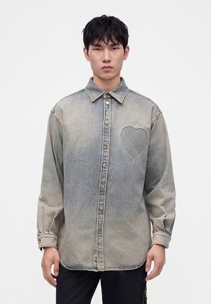 Jeune homme portant une chemise en jean gris délavé à boutons avec un détail cœur brodé sur le côté gauche de la poitrine, debout devant un fond blanc uni.