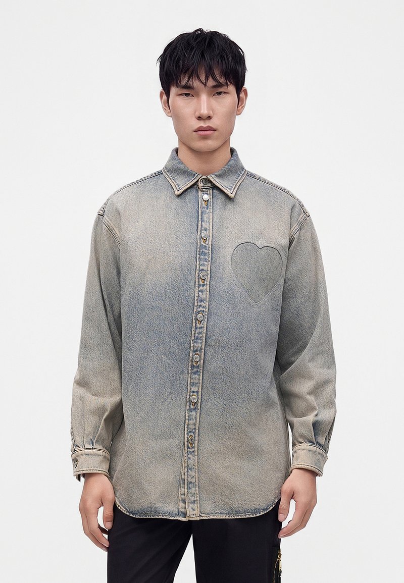 Jeune homme portant une chemise en jean gris délavé à boutons avec un détail cœur brodé sur le côté gauche de la poitrine, debout devant un fond blanc uni.