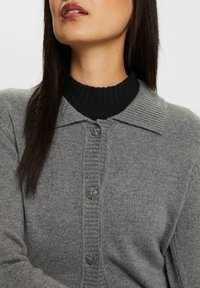 Šedý pletený cardigan s žebrovaným černým límcem. Obsahuje šest šedých knoflíků na přední straně a má jemnou texturu. Zvýrazněn vertikálními liniemi.