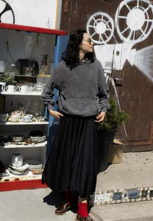 Szara bluza z polaru z kieszenią z przodu, zestawiona z długą czarną spódnicą. Widoczne brązowe buty i czerwone skarpetki. W tle półki z naczyniami.