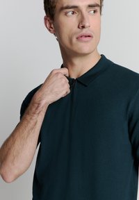 Donkergroen, met rits polo-shirt met een kraag. Bevat een gebreide stof en korte mouwen. Minimalistisch ontwerp zonder zichtbare patronen.