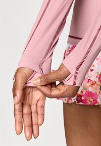 Top lung cu mâneci lungi, roz deschis, cu găuri pentru degete și cusături în contrast de culoare roz închis; pantaloni scurți cu model floral vizibili la margine.