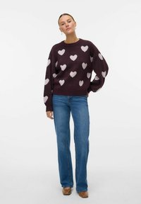 Maglione lavorato a maglia bordeaux con ampi motivi di cuori bianchi, vestibilità rilassata, colletto a coste, abbinato a jeans blu chiaro a zampa d'elefante e scarpe marroni.