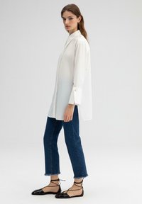 Witte blouse met lange mouwen en een aansluitende kraag, zijsplitten en knoopmanchetten, gecombineerd met een blauwe cropped jeans en zwarte veterschoenen.