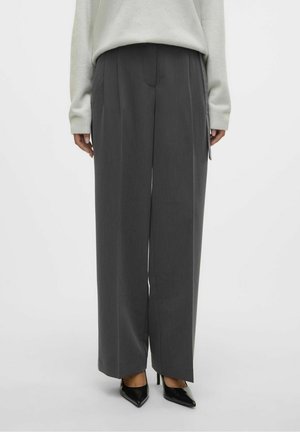 Person trägt weit geschnittene dunkelgraue Hose, hellfarbenen Pullover und schwarze spitze High Heels, steht vor einfarbigem Hintergrund.